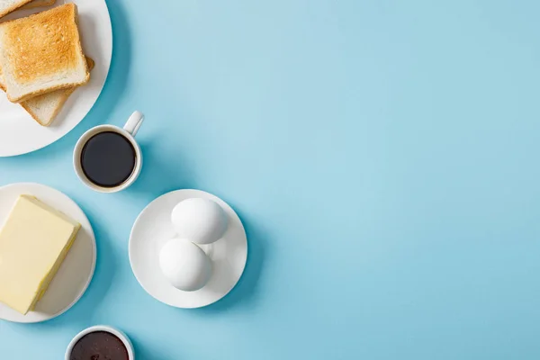 Вид зверху на свіжі яйця, варення, масло, каву і два тости на білих тарілках на синьому фоні — стокове фото
