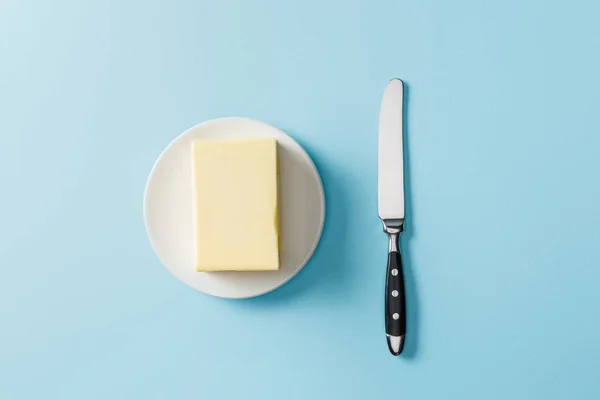 Vue du dessus du beurre sur plaque blanche et couteau sur fond bleu — Photo de stock