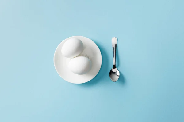 Vista superior de cucharadita cerca de huevos cocidos en plato blanco sobre fondo azul - foto de stock