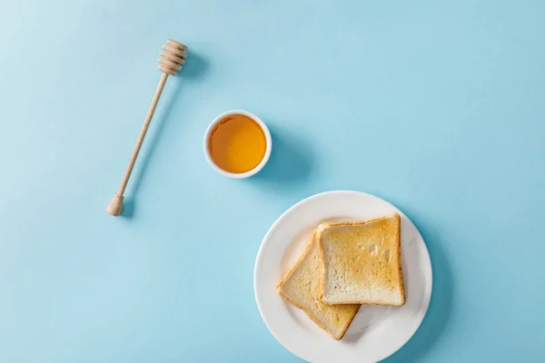 Top view of honey in bowl, wooden dipper and two toasts on white plate on blue background — Stock Photo