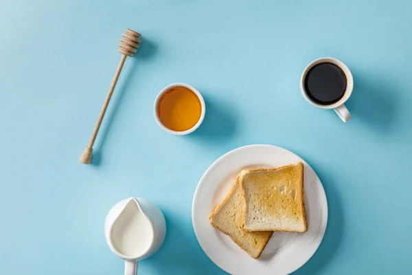 Top view of honey in bowl, wooden dipper, coffee, milk and two toasts on white plate on blue background — Stock Photo