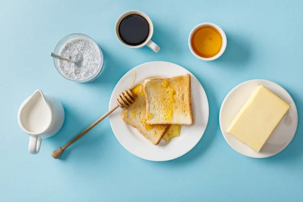 Top view of coffee, milk, yogurt with chia seeds, butter, honey, toasts with honey and wooden dipper on white plates on blue background — Stock Photo