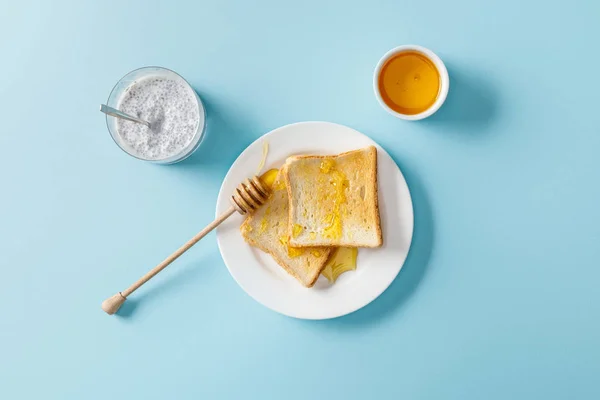 Top view of yogurt with chia seeds, toasts with honey and honey dipper on white plate on blue background — Stock Photo