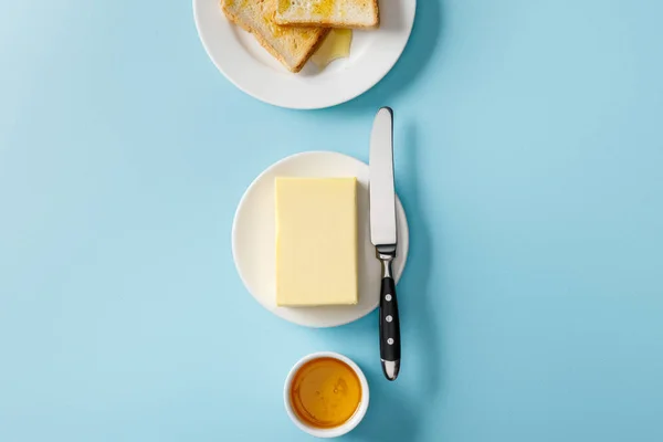 Vista superior de mantequilla, cuchillo, tostadas en platos blancos y tazón con miel sobre fondo azul - foto de stock