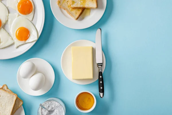 Vista superior de mantequilla, tostadas, huevos frescos y fritos en platos blancos, yogur con semillas de chía y tazón con miel sobre fondo azul - foto de stock