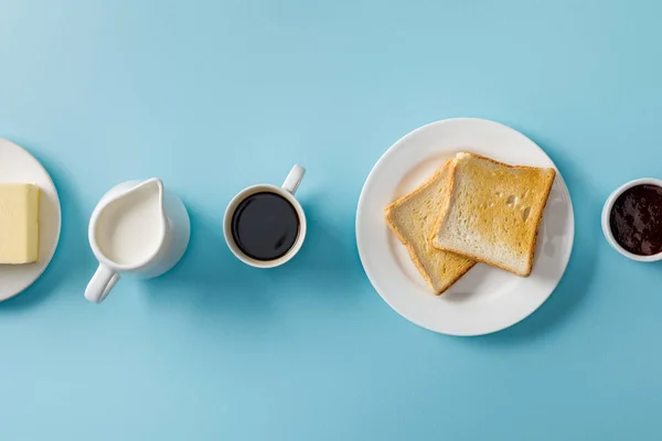 Draufsicht auf Tasse Kaffee, Milch, Butter, Marmelade und zwei Toasts auf weißem Teller auf blauem Hintergrund — Stockfoto