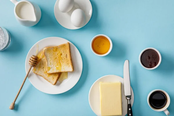 Top view of served breakfast with yogurt, milk, coffee, jam, honey, butter and knife, toasts on white plates on blue background — Stock Photo