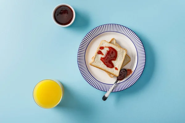 Vista superior de jugo de naranja, tostadas, tazón y cuchara con mermelada en el plato sobre fondo azul - foto de stock