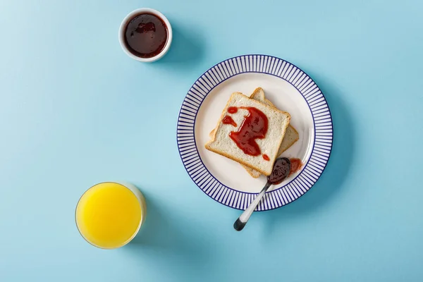 Vue du dessus du jus d'orange, des toasts, du bol et de la cuillère avec confiture sur fond bleu — Photo de stock