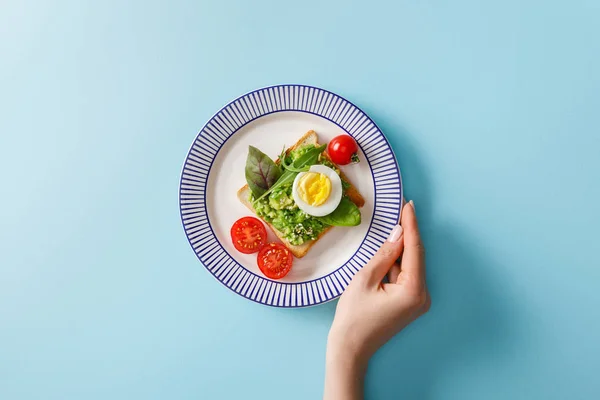 Cropped view of woman holding ornamental plate with guacamole, boiled egg and greenery on toast near cherry tomatoes on blue background — Stock Photo