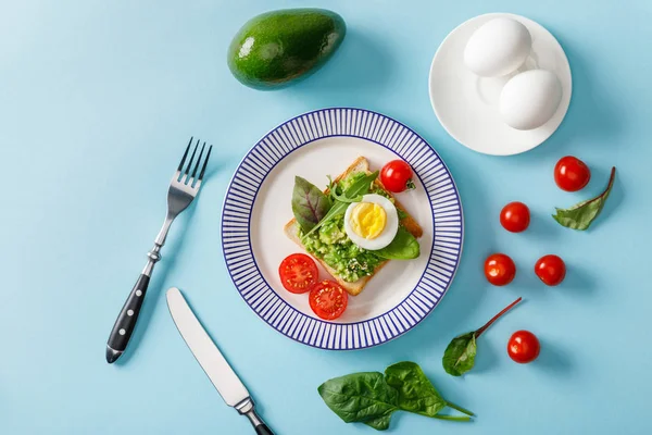 Top view of toast with guacamole, avocado, boiled eggs, scattered cherry tomatoes and spinach on blue background — Stock Photo