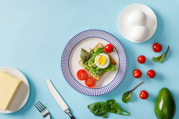 Тосты с гуакамоле, авокадо, вареные яйца, масло, разбросанные помидоры черри и шпинат на голубом фоне — стоковое фото
