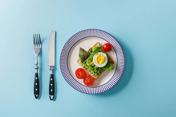 Top view of toast with guacamole, boiled egg cherry tomatoes, fork and knife on blue background — Stock Photo