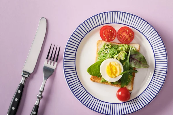 Top view of toast with guacamole, boiled egg, spinach, cherry tomatoes, fork and knife on ornamental plate on violet background — Stock Photo