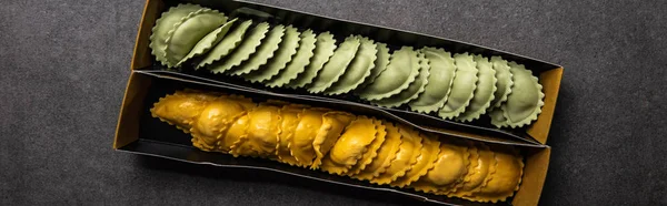 Top view of green and yellow raw ravioli in cardboard box on grey textured surface, panoramic shot — Stock Photo