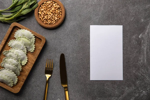 Servi sur une planche de bois raviolis verts avec farine près d'un bol avec graines, herbes, couverts et carte vide — Photo de stock