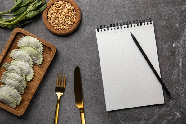 Servi sur une planche de bois raviolis verts avec farine près d'un bol avec graines, herbes, couverts et cahier vierge avec crayon — Photo de stock