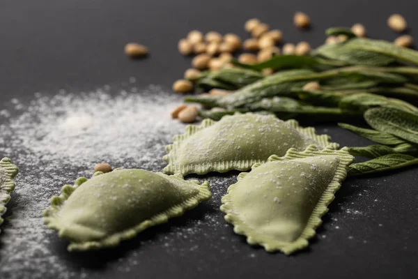 Selective focus of green ravioli, sage and scattered flour and pine nuts on black table — Stock Photo