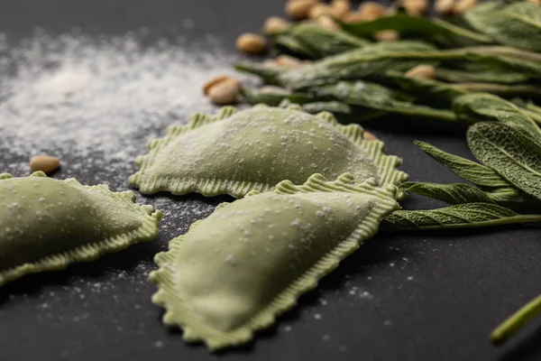 Selektiver Fokus grüner Ravioli in der Nähe von Salbei, Mehl und Pinienkernen auf schwarzem Tisch — Stockfoto