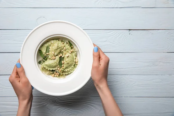 Cropped view of woman holding plate with green ravioli with pine nuts and sage at wooden table — Stock Photo