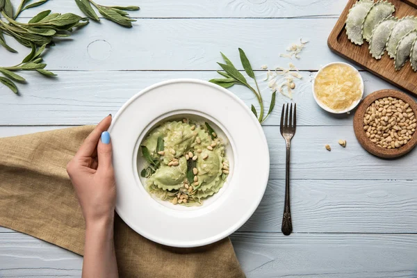 Cropped view of woman holding plate with green ravioli, pine nuts, sage near grated cheese on white wooden table with fork and sackcloth — Stock Photo