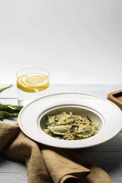 Grüne Ravioli mit Pinienkernen und Salbei am Holztisch mit Serviette und Wasser mit Zitrone isoliert auf grau — Stockfoto