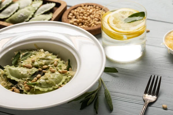 Vista de perto de ravioli verde com pinhões, queijo ralado e sálvia à mesa de madeira perto da água com limão — Fotografia de Stock