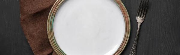 Top view of vintage white empty round plate on black wooden table near brown napkin and silver fork, panoramic shot — Stock Photo