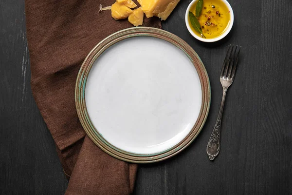 Draufsicht auf weißen, leeren runden Teller auf schwarzem Holztisch neben brauner Serviette, Käse, Öl mit Gewürzen und Silbergabel — Stockfoto