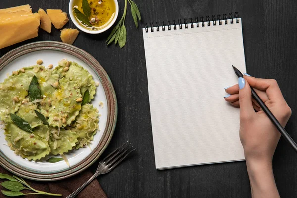 Vista cortada de mulher escrevendo em caderno em branco com lápis perto servido ravioli verde — Fotografia de Stock