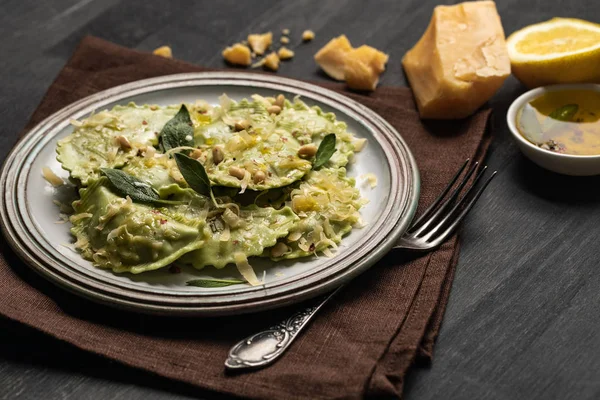 Deliziosi ravioli verdi con salvia, formaggio e pinoli serviti su tavolo di legno nero con forchetta, limone e tovagliolo — Foto stock