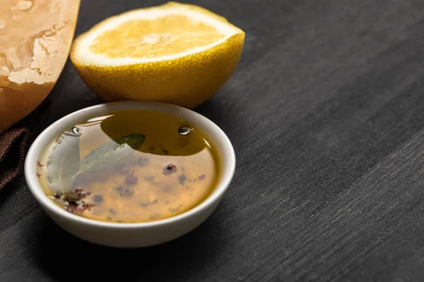 Vue rapprochée de l'huile aromatique dans un bol avec des épices et des herbes près du citron et du fromage sur une table noire en bois — Stock Photo