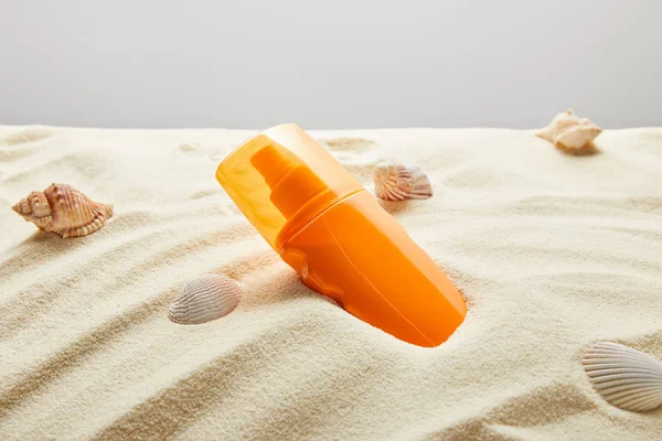 Sonnencreme in orangefarbener Flasche in Sand mit Muscheln auf grauem Hintergrund — Stockfoto