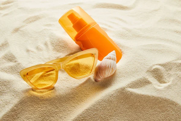 Gafas de sol de estilo amarillo y protector solar en botella naranja sobre arena con concha - foto de stock