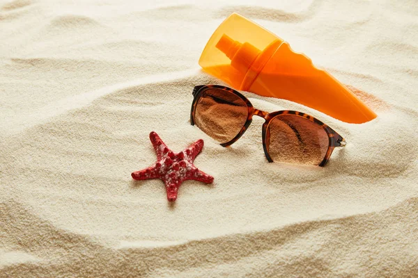 Коричневі стильні сонцезахисні окуляри на піску з червоною зіркою і сонцезахисним кремом в помаранчевій пляшці — стокове фото