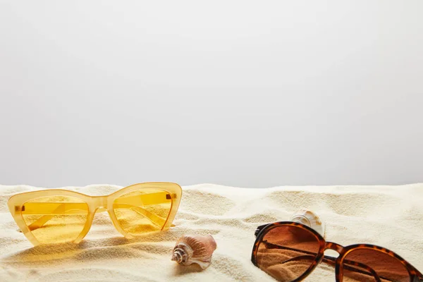 Yellow and brown stylish sunglasses on sand with seashells on grey background — Stock Photo