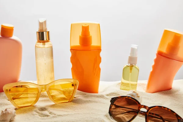 Vários produtos de proteção solar em garrafas na areia perto de óculos de sol na moda e conchas no fundo cinza — Fotografia de Stock