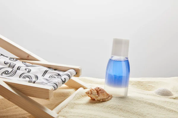 Lotion bleue en bouteille dans le sable près des coquillages et chaise longue sur fond gris avec espace de copie — Photo de stock