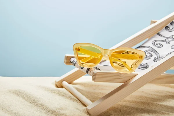 Gafas de sol amarillas y silla de cubierta sobre arena sobre fondo azul - foto de stock