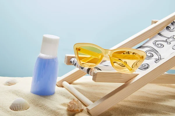 Синій лосьйон у піску біля черепашок, жовті сонцезахисні окуляри та стілець на синьому фоні — стокове фото