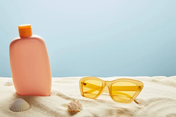 Lotion solaire dans le sable près des coquillages et des lunettes de soleil jaunes sur fond bleu — Photo de stock