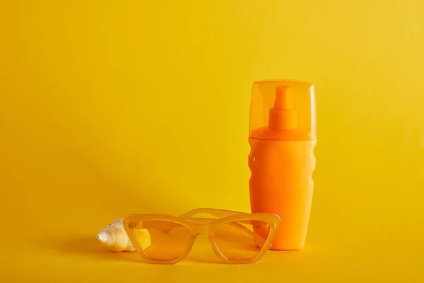 Sonnencreme in orangefarbener Flasche in der Nähe von Sonnenbrille und Muschel auf dunkelgelbem Hintergrund — Stockfoto