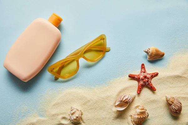 Vue du dessus de la crème solaire en bouteille près des coquillages, étoiles de mer, sable et lunettes de soleil sur fond bleu — Photo de stock