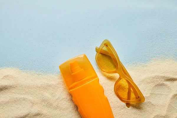 Vista superior do protetor solar em garrafa de laranja perto de óculos de sol no fundo azul com areia — Fotografia de Stock