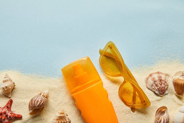 Vue du dessus de la crème solaire en bouteille orange près des lunettes de soleil sur fond bleu avec sable et coquillages — Photo de stock