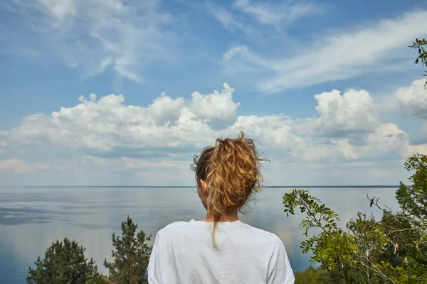 Back view of young woman looking at landscape with green leafy trees, river and blue sky — Stock Photo