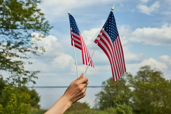 Cropped view of young woman holding two small flags in hand on river and forest background — Stock Photo