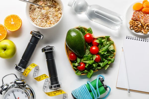 Vista dall'alto di attrezzature sportive, nastro di misurazione, sveglia e notebook vicino al cibo dieta e acqua su sfondo bianco — Foto stock