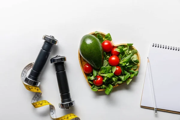 Vista dall'alto di verdure in ciotola a forma di cuore, metro a nastro, notebook e manubri su sfondo bianco — Foto stock