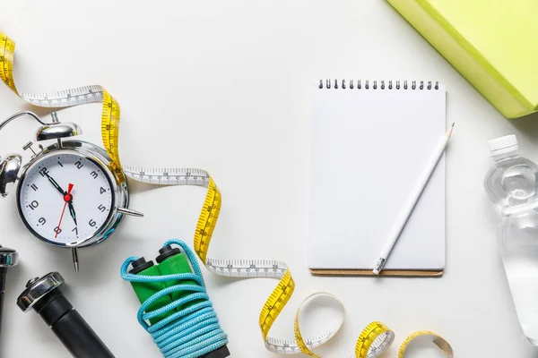 Top view of sport equipment, measuring tape, alarm clock, water and blank notebook on white background — Stock Photo